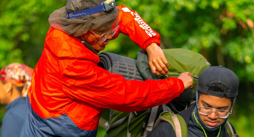 One person adjusts the backpack of another person while outdoors